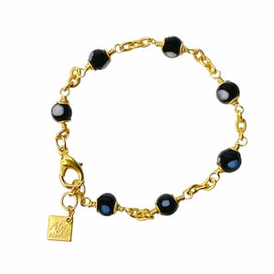 May include: Gold chain bracelet with black and white glass beads. The bracelet has a clasp closure and a small square tag with the text "Ali by Alzima".