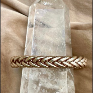 May include: A braided gold-toned metal bracelet resting on a clear quartz crystal. The bracelet has a woven design.