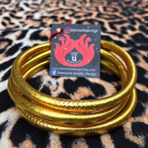 May include: Gold-colored bangles stacked on a leopard print fabric. A tag with a red flame design and the text "Lucky for U" is attached. The tag also includes social media handles and a website address.