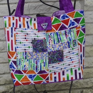 May include: A colorful tote bag with a patchwork design. The bag features a variety of geometric patterns in shades of purple, orange, green, and blue. It has a purple handle and a matching clasp. The bag is made of fabric and appears to be handmade.