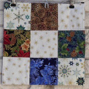 May include: A patchwork fabric square with nine distinct sections. The sections feature various holiday-themed patterns, including snowflakes, pinecones, and floral designs in colors like white, gold, blue, red, and green. The fabric is held by a metal clip.