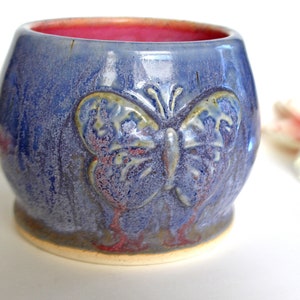 Op de afbeelding: Een kleine, blauwe keramische pot met een reliëf van een vlinder. De pot heeft een rode rand en een witte basis.