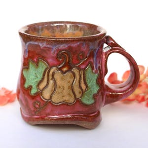 May include: A ceramic mug with a glossy red and purple glaze featuring a raised, tan-colored pumpkin design in the center. The pumpkin is flanked by two green leaves, and the mug has a curved handle on the right side with a wide, rounded base.
