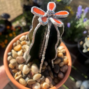 May include: A stained glass cactus with a red flower in a terracotta pot filled with small pebbles. The cactus has a dark green body and silver-colored metal accents. The flower has red petals with silver outlines.