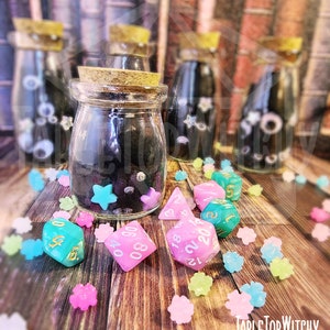 May include: A clear glass jar with a cork lid, filled with black material and decorated with blue stars and pink gems. Several dice in pink and teal are scattered around the jar, along with colorful star-shaped candies. The text "TableTop Witchy" is visible.