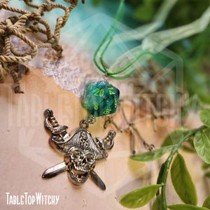 May include: A pirate-themed pendant with a skull and crossed swords charm, paired with a green and clear D20 die. The pendant is suspended from a green cord. The text "TableTop Witchy" is visible at the bottom.