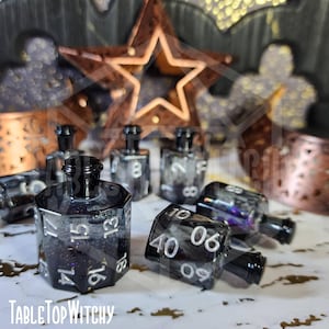 May include: Black and clear resin dice with white numbering, resembling potion bottles. The dice are scattered on a white surface, with a copper star and decorative metal cutouts in the background. The text "TableTop Witchy" is visible.
