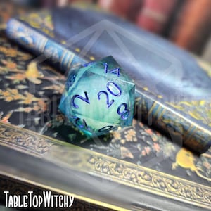 May include: A large, translucent blue and green twenty-sided die with dark blue numbering. A matching pen rests behind the die. The text "TableTopWitchy" is visible at the bottom of the image.