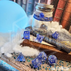May include: A clear glass jar with a cork stopper, adorned with silver and blue ribbons and a blue bead. Several blue, translucent dice with white numbering are scattered around the jar. The text "Tabletop Witchy" is visible in the bottom right corner.