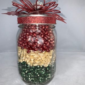 May include: A clear glass mason jar filled with layers of red, white, and green kernels. The jar is topped with a red glittered lid and a decorative red bow. The jar is sitting on a white surface.