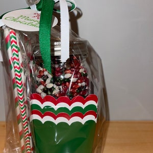 May include: A clear plastic bag filled with a glass jar of red, green, and white candy, a green and white striped paper straw, a red and white striped paper straw, and a green paper cup with red and white scalloped edges. The bag is tied with a green and white ribbon and a tag that says "Merry Christmas".