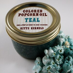 May include: A glass jar filled with teal coloured popcorn oil. The jar has a gold lid with a label that reads "COLORED POPCORN OIL TEAL Add a POP of Color to your occasion KITTS KERNELS". Teal popcorn is scattered around the jar.
