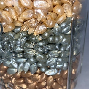 May include: A glass jar filled with three layers of colorful popcorn kernels. The top layer is gold, the middle layer is silver, and the bottom layer is gold.