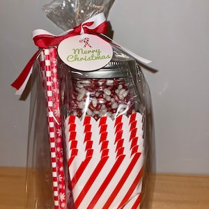 May include: A clear plastic bag filled with a red and white striped popcorn container, a glass jar filled with red and white popcorn kernels, and two red and white striped straws. A white tag with the text "Merry Christmas" is attached to the bag.