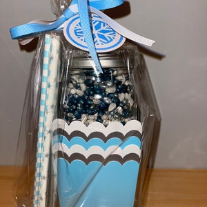 May include: A clear plastic bag filled with a blue and silver snowflake themed gift. The gift includes a glass jar filled with blue and silver sprinkles, a blue and white striped paper straw, and a blue and white checkered paper napkin. The bag is tied with a blue and white ribbon and a snowflake tag.