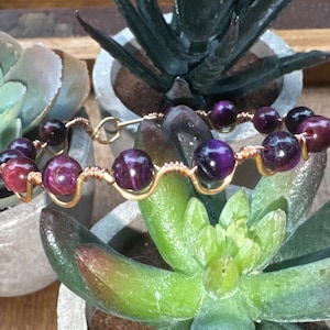 May include: A bracelet featuring round, polished purple and deep red beads connected by a continuous, wavy gold-toned wire. The wire is wrapped around each bead, creating a structured, linked design that secures the stones in a repeating pattern.