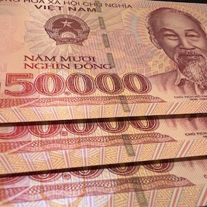 Puede incluir: Primer plano de varios billetes vietnamitas de 50.000 Dong. Los billetes son predominantemente melocot&oacute;n y rojos, con un retrato de Ho Chi Minh y el emblema vietnamita. El texto "Năm Mươi Ngh&igrave;n Đồng" es visible.
