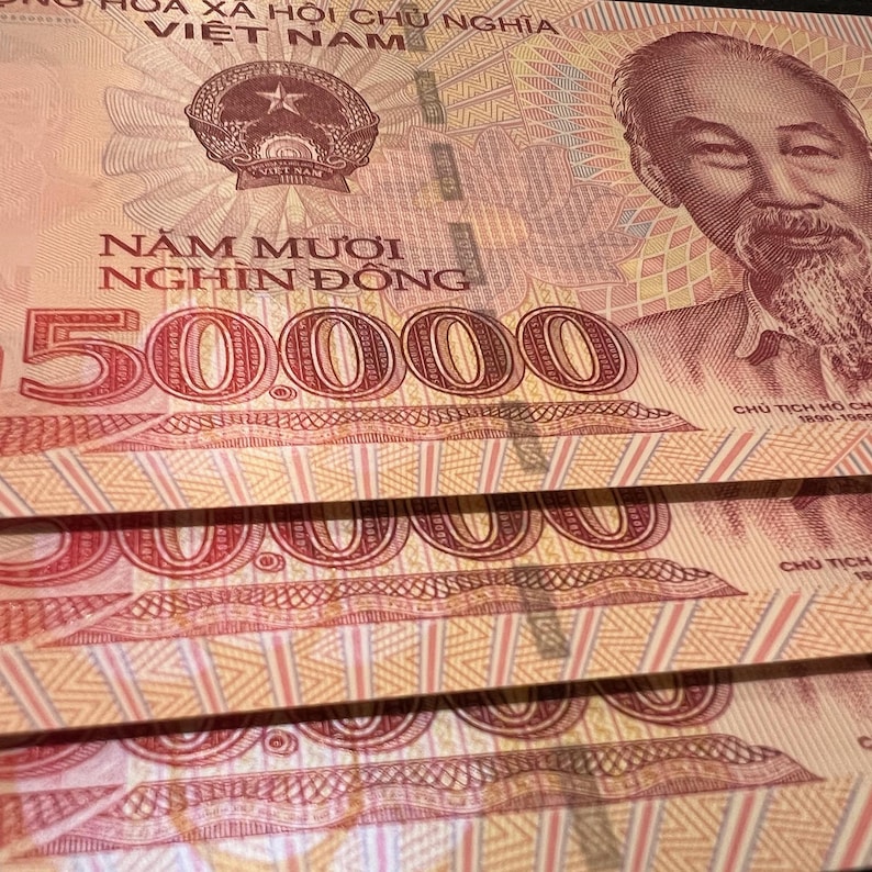Puede incluir: Primer plano de varios billetes vietnamitas de 50.000 Dong. Los billetes son predominantemente melocot&oacute;n y rojos, con un retrato de Ho Chi Minh y el emblema vietnamita. El texto "Năm Mươi Ngh&igrave;n Đồng" es visible.