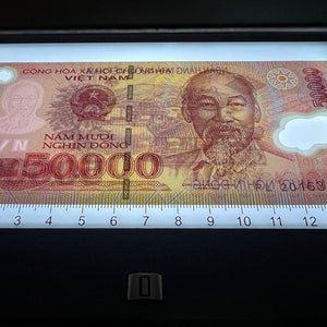 Puede incluir: Un billete vietnamita de 50.000 Dong, con un retrato de Ho Chi Minh. El billete tiene texto en vietnamita y el n&uacute;mero 50.000 se muestra de forma destacada. El billete est&aacute; iluminado en una mesa de luz.
