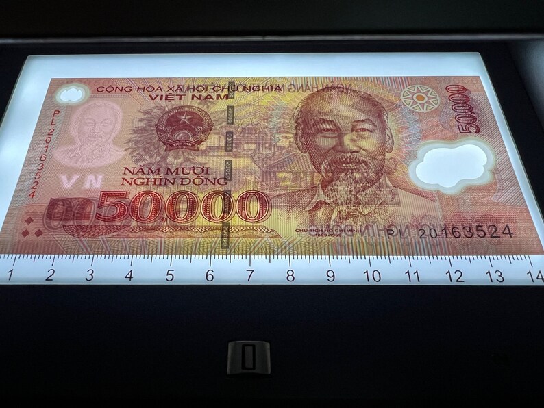 Puede incluir: Un billete vietnamita de 50.000 Dong, con un retrato de Ho Chi Minh. El billete tiene texto en vietnamita y el n&uacute;mero 50.000 se muestra de forma destacada. El billete est&aacute; iluminado en una mesa de luz.
