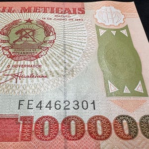May include: A close-up of a Mozambique 100,000 Meticais banknote. The bill features text in Portuguese, a coat of arms, and the serial number FE4462301. The note has a green and red color scheme.