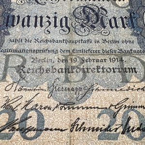 May include: An antique German 20 Mark banknote from 1914, featuring ornate floral patterns and text in German. The note has a faded blue and beige color scheme, with the text "wanzig Mark" prominently displayed.