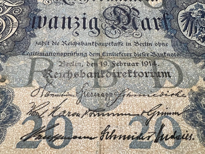 May include: An antique German 20 Mark banknote from 1914, featuring ornate floral patterns and text in German. The note has a faded blue and beige color scheme, with the text "wanzig Mark" prominently displayed.