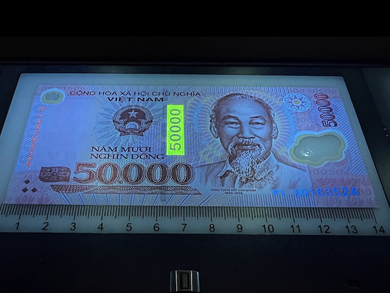 Puede incluir: Un billete vietnamita de 50.000 Dong bajo luz ultravioleta. El billete presenta un retrato de Ho Chi Minh y el texto "CỘNG H&Ograve;A X&Atilde; HỘI CHỦ NGHĨA VIET NAM". Una regla se coloca debajo, con medidas en cent&iacute;metros.