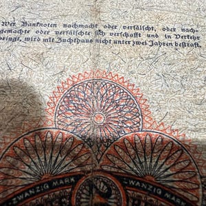May include: Close-up of a vintage German 20 Mark banknote. The note features intricate circular designs in red, black, and orange. Text in German is visible at the top, with the words "ZWANZIG MARK" at the bottom.
