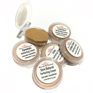 May include: A collection of small, round cosmetic containers with light beige concealer foundation. The containers are labeled with names like "Cashmere" and "Porcelain." The text "Minerals By Sandrah" and "Perfecting Cream" are also visible.