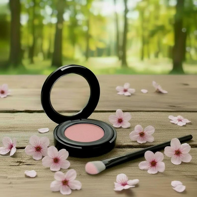 May include: A round black compact containing soft pink powder blush with a hinged lid, accompanied by a black makeup brush with a light-colored tip. The items are surrounded by small, delicate light pink cherry blossom flowers.