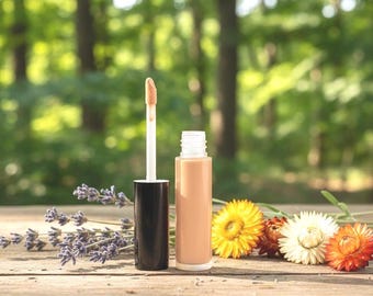 May include: A liquid concealer in a clear cylindrical tube with a black cap and an attached applicator wand. The product is light beige in colour. Beside the container are sprigs of purple lavender and three dried flowers in yellow, white, and orange.