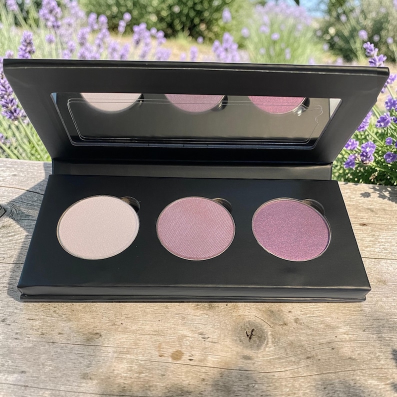 May include: A black eyeshadow palette with three circular pans of light pink, medium pink, and purple eyeshadow. The palette has a built-in mirror and is open, revealing the colors. The background features lavender flowers and a wooden surface.