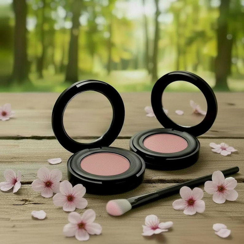 May include: Two round black compacts containing pressed pink powder blush, accompanied by a black makeup brush with a soft tip. The items are arranged among several small, light pink five-petaled flowers on a weathered wooden surface.
