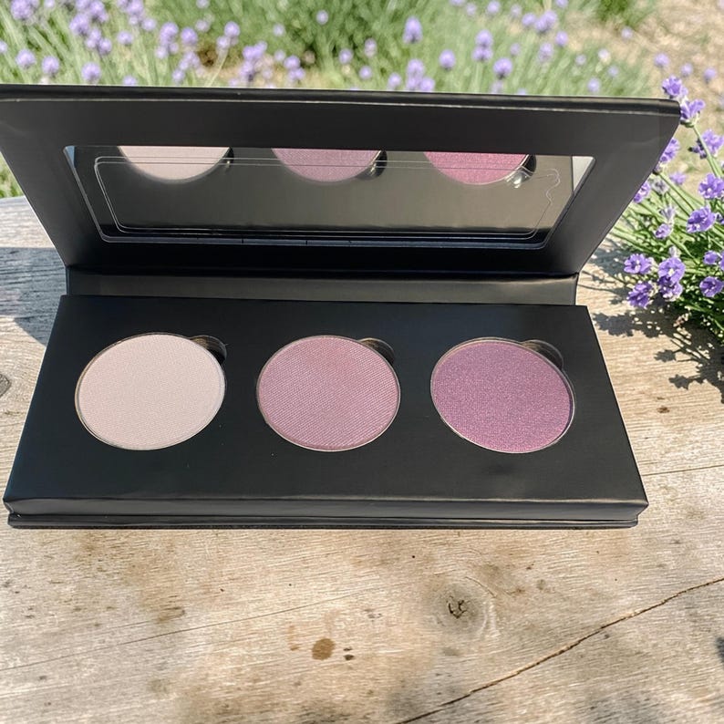 May include: A black eyeshadow palette with three circular pans of varying shades of pink and purple. The palette has a built-in mirror in the lid. The palette is open, revealing the colors, and is set on a wooden surface.