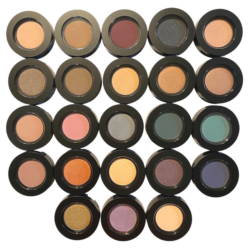 May include: A collection of twenty-three round eyeshadow pots arranged in a grid, each featuring a black plastic casing and a unique pressed powder color, including shades of beige, brown, pink, orange, yellow, green, blue, and purple.