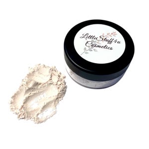 May include: A small, round, clear cosmetic jar with a black lid featuring a white label that reads Little Stuff 4u Cosmetics with a leaf illustration. Beside the jar is a small pile of fine, light beige loose powder with a soft, matte texture.