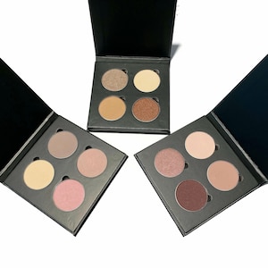 May include: Three black eyeshadow palettes, each with four circular pans of eyeshadow. The palettes are open and show a variety of colors, including brown, beige, pink, and purple.