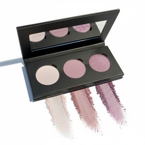 May include: A black eyeshadow palette with three circular pans in light pink, medium pink, and shimmering purple. Swatches of each color are displayed below the pans. A mirror is in the lid.