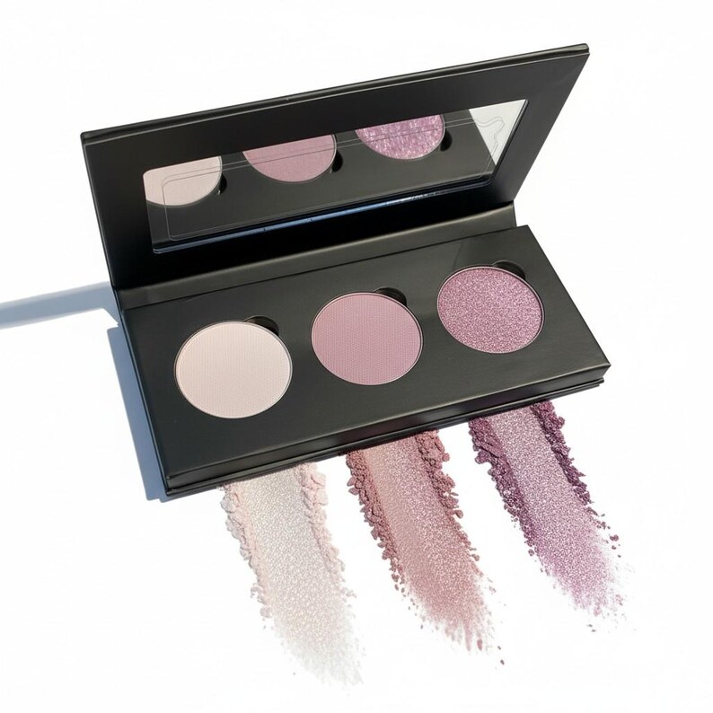 May include: A black eyeshadow palette with three circular pans in light pink, medium pink, and shimmering purple. Swatches of each color are displayed below the pans. A mirror is in the lid.