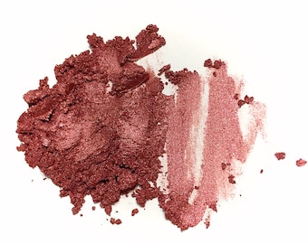 May include: A pile of loose, shimmering dark rose-colored pigment powder next to a smooth, lighter pink metallic swatch. The texture appears fine and granular, with a reflective, pearlescent finish visible across both the concentrated mound and the streak.
