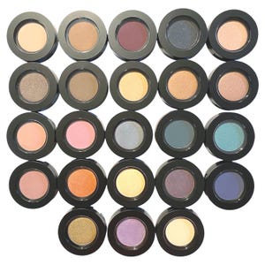 May include: A collection of twenty-two individual round eyeshadow pots arranged in a grid. Each container features a black circular frame holding a pressed powder pigment in various shades including beige, brown, pink, blue, purple, and gold.