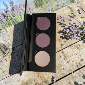 May include: A black eyeshadow palette with three circular pans in shades of purple and mauve. The palette is open, revealing the colors against a wooden surface with lavender flowers in the background. The eyeshadows are in a compact, portable design.