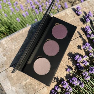 May include: A black eyeshadow palette with three circular pans of varying shades of purple and mauve. The palette is open, revealing the colors against a wooden surface, with lavender flowers in the background.