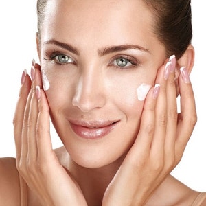 May include: A face with light skin tone and green eyes, featuring small dabs of white facial cream applied to the cheeks. The skin appears smooth and hydrated, with a subtle sheen. The facial expression is neutral with a soft, closed-mouth smile.