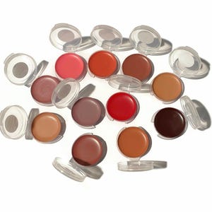 May include: A collection of 14 round lip balm pots with various shades of red, pink, brown, and nude. The pots are clear plastic with a silver lid.