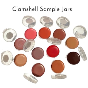 May include: A collection of round, clear plastic clamshell sample jars containing various shades of cream-based cosmetic products in colors ranging from light beige and pink to deep red, brown, and dark plum. The text Clamshell Sample Jars is visible.