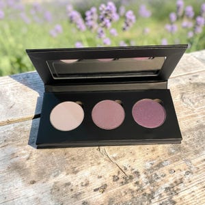 May include: A black eyeshadow palette with three circular pans of color. The shades range from light beige to deep purple. The palette has a built-in mirror and is open, revealing the colors. The background is blurred, showing lavender flowers.