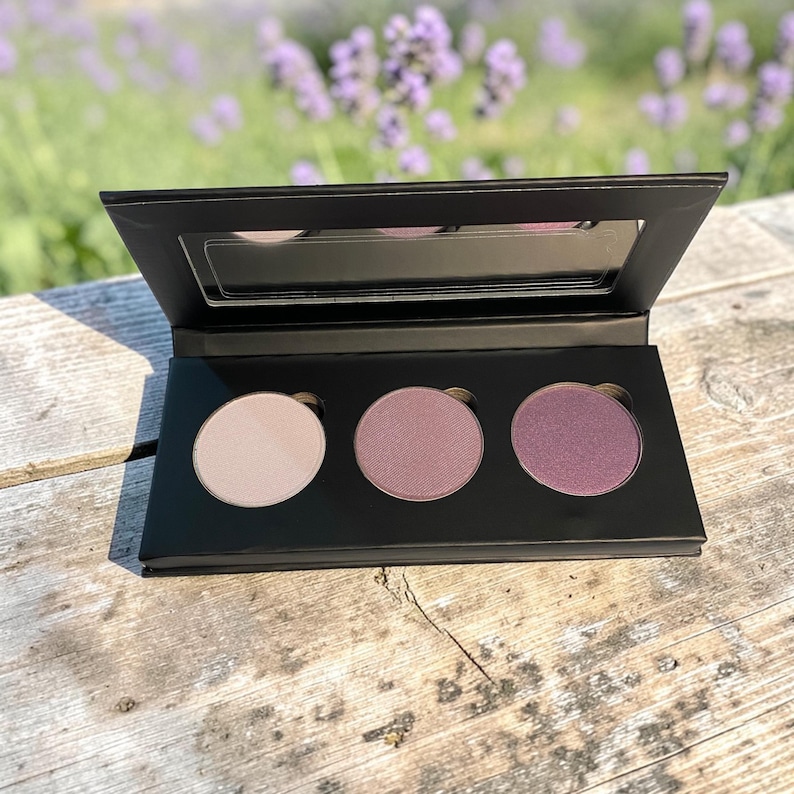 May include: A black eyeshadow palette with three circular pans of color. The shades range from light beige to deep purple. The palette has a built-in mirror and is open, revealing the colors. The background is blurred, showing lavender flowers.