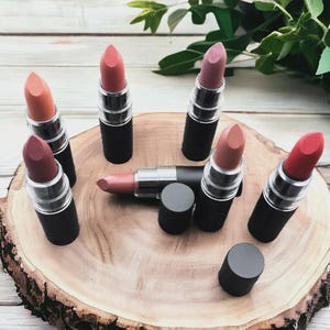 Pode incluir: Uma variedade de batons em tons de rosa, p&ecirc;ssego e vermelho, dispostos sobre uma superf&iacute;cie de madeira. Os batons t&ecirc;m tubos pretos com detalhes prateados e tampas transparentes. Um batom est&aacute; destampado, revelando sua cor.